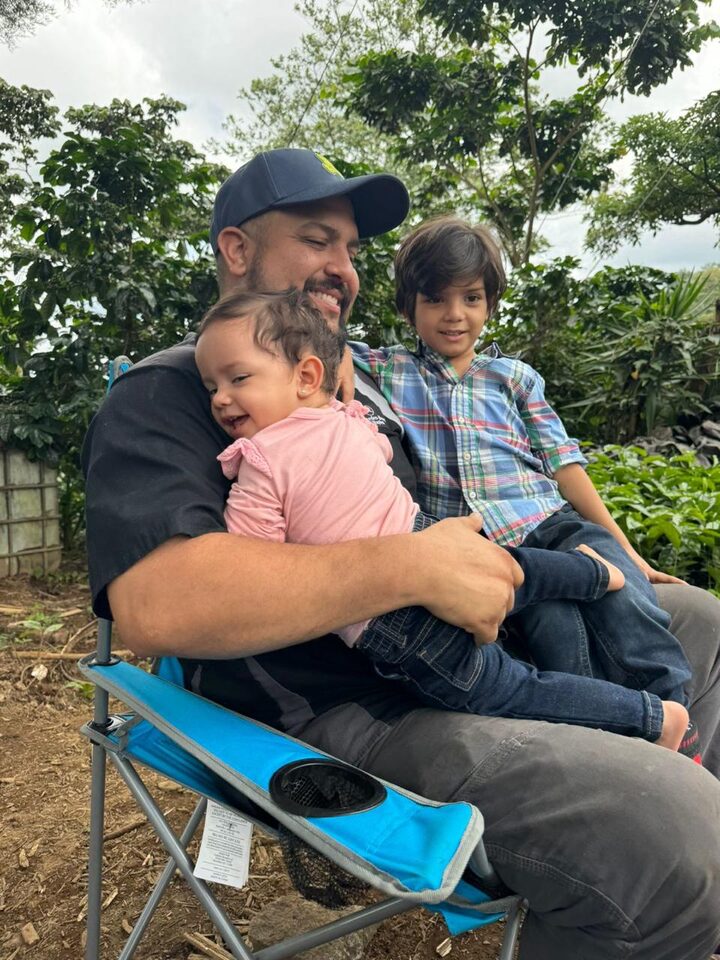 Guillermo Ríos sentado al aire libre entre árboles, con dos niños pequeños en brazos.